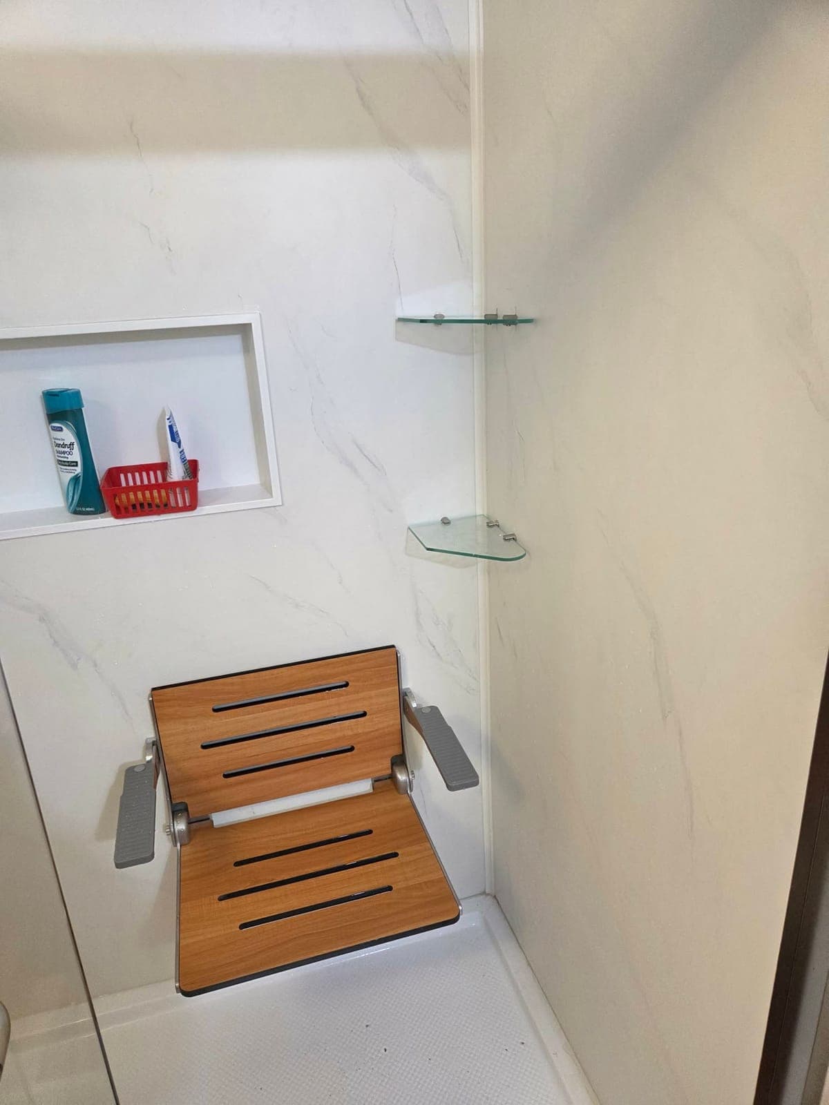 Shower interior with marble-look wall panels, corner glass shelves, a recessed niche, and a folding teak shower seat